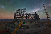 Standing tall, and appearing in shape much like the ruins of the Roman colosseum, the ELT dominates the centre of this image. Its concrete circular base is topped by a criss-crossed network of steel beams. The image is taken at night time and behind the ELT dome, which is still under construction, the sky is alive with stars. The horizon has a red hue to it, while greens and blues dance above. Over to the right of the dazzling sky, the bright white of the Milky Way is streaking across the sky. A tall crane is also present over on the right hand side of the image, while another is peeking out from the bottom left.