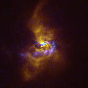 The background of this image is dark, but in its centre lurks a swirling ghostly figure, which extends towards the edge of the picture. At the very centre there is a small bright region and erupting out of it there is a poorly defined, fuzzy edged cloud and blobs of material in yellow and blue, respectively. The yellow cloud extends far out in the image, making an elongated spiral shape that gets dimmer and less defined as it reaches the top and bottom of the frame. Meanwhile, the blue blobs only extend downwards from the centre and to a fraction of the distance of the yellow spiral cloud. The blobs twist away from the central bright region, forming a tight U-shape lying on its right side.