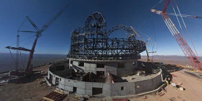 The image shows the ELT construction site in the Atacama desert, kissed by the white moonlight that fills the air. Although the Moon itself isn’t in the frame, you can feel its presence nearby. The night sky is a deep blue, becoming slightly whiter as it meets the brown horizon of the desert. Dominating the centre of the image is the giant ELT structure under construction, with its circular concrete base at the bottom and its magnificent steel-framed dome on top. The criss-cross steel frame now has two complete arches at the top. On the right and left of the ELT are two cranes leaning in towards the telescope.